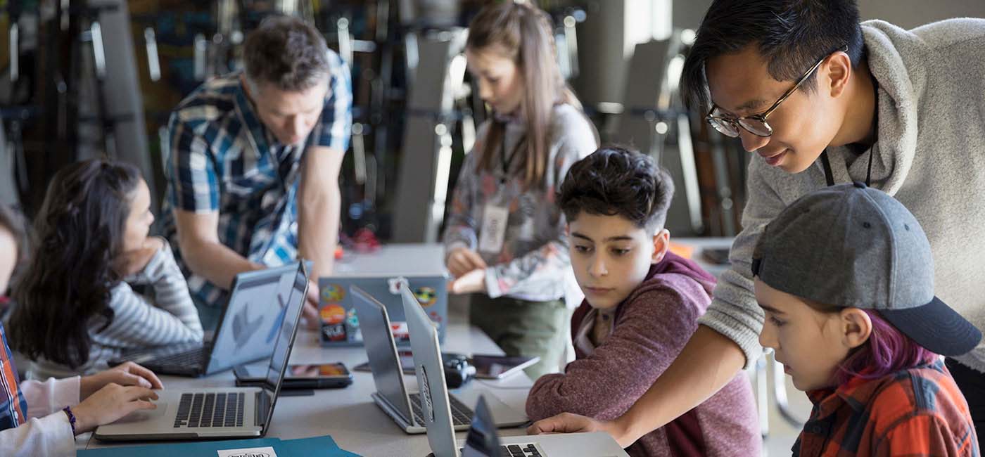 A photo of five children using laptops, sitting at a rectangle table in a classroom,  with two male teachers helping them!''