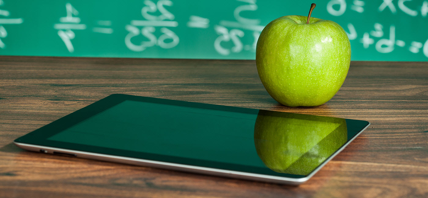 Ipad on a desk with an apple and chalkboard in the background.